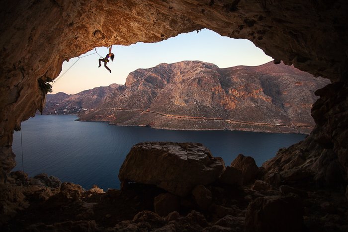 a rockclimber above a cave by water and a mountainous landscape - adventure photography skills
