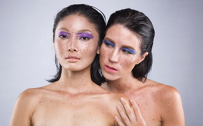 Artistic nude photography of two naked female models with purple eyeliner and emphasized freckles 