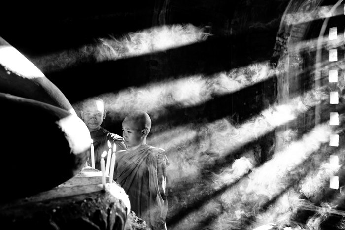 Atmospheric monochrome shot of young monks lighting candles with incident light shining through a doorway