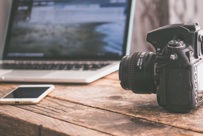 Photo of a laptop, a camera, and a phone on a wooden desk