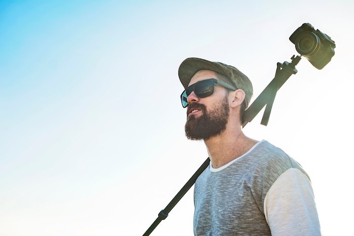 A man with a beard, sunglass, hat, and a monopod with a camera resting on his shoulder