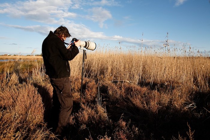 A wildlife photographer using a monopod with a camera and lens in a field with tall grass