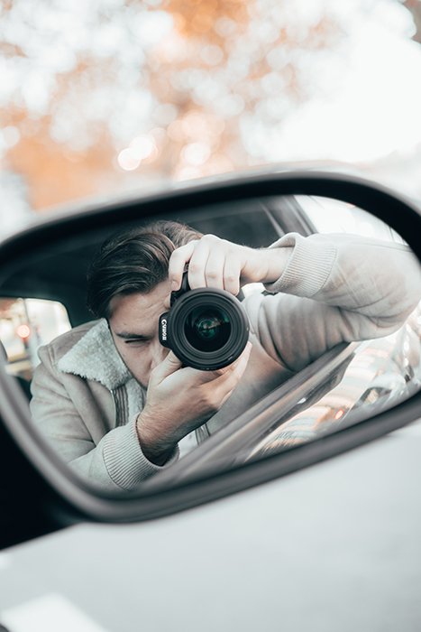 Reflection in a car wing mirror showing a man holding a camera 