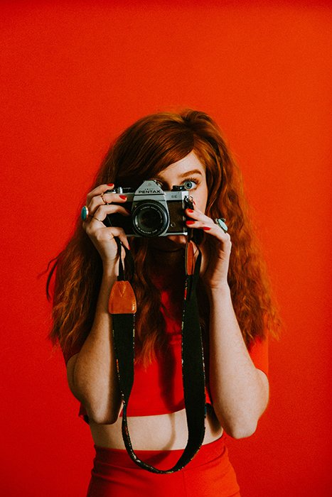 Red haired girl in front of a bright red screen holding a Pentax camera 