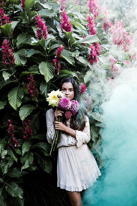 A girl standing and holding flowers, a large billow of smoke is coming towards her from the right hand side 