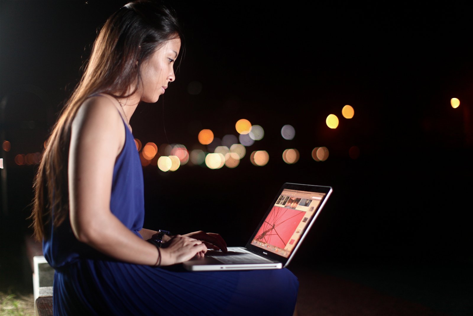 A woman using a laptop outside at night with bokeh lights in the distance
