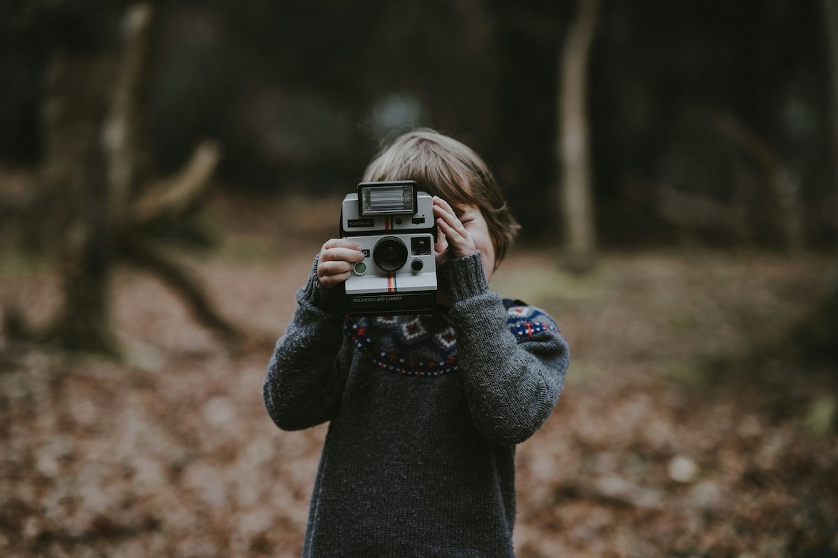 A boy holding up a Polaroid camera in the forest to show photography for kids