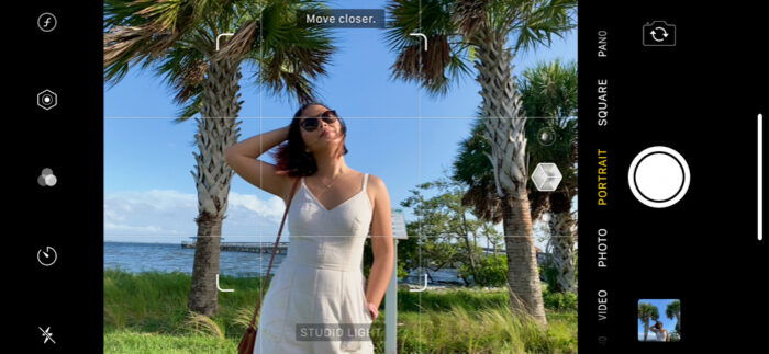 Screenshot of an iPhone's camera app in Portrait mode taking a picture of a woman posing in a white dress with palm trees in the background