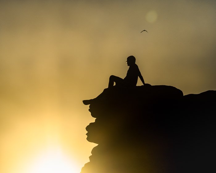 Silhouette of a boy sitting on the edge of a cliff