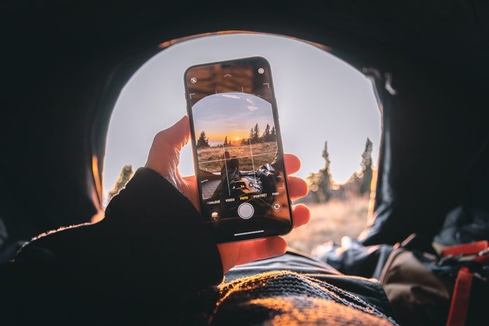 A person's hand holding up a smartphone with the camera app showing a setting sun landscape outside a tent