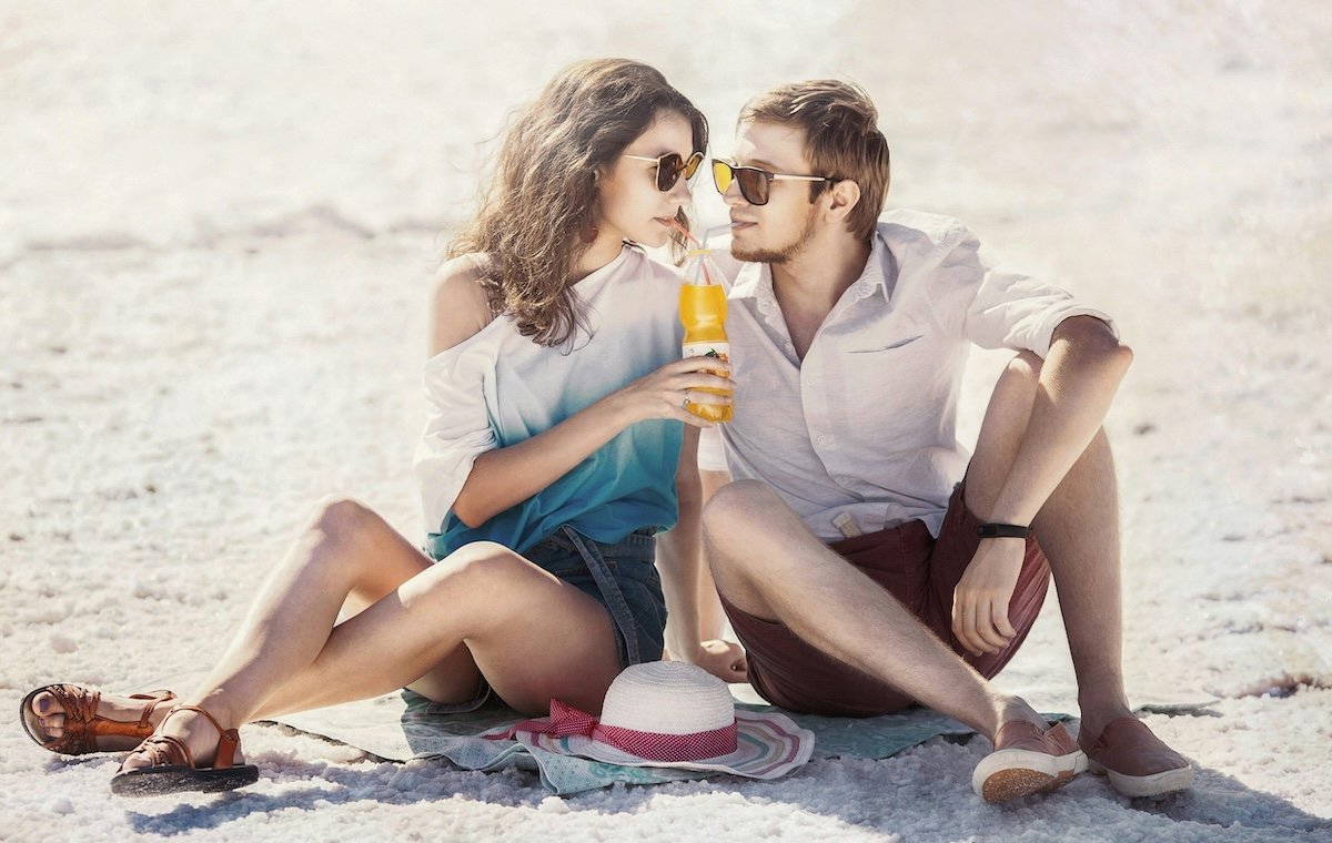 Two people looking at each other over a drink sitting on a beach for couple poses