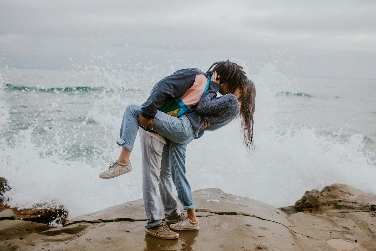Two people kissing with a wave washing ashore and the man tilting her back and lifting the woman's leg for couple poses