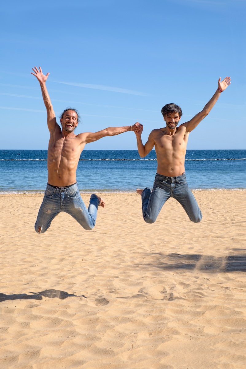 Two men in jeans and no shirt jumping with hands held on a beach for couple poses