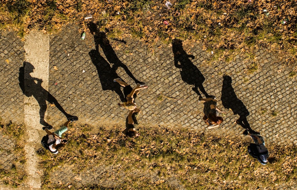 Overhead shot of people walking on a stone and grass walkway with their harsh black shadows cast on the ground