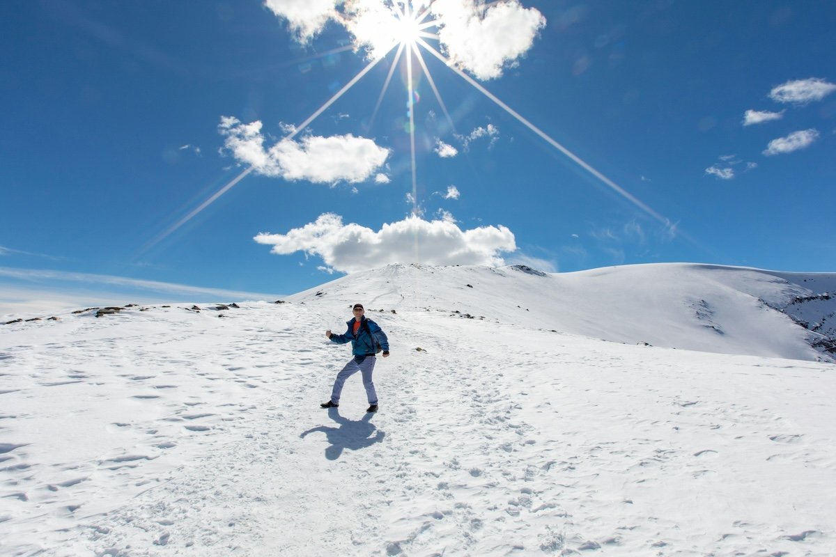 A person posing on top of a mountain plateau covered with snow and the sun behind with some white clouds and blue sky