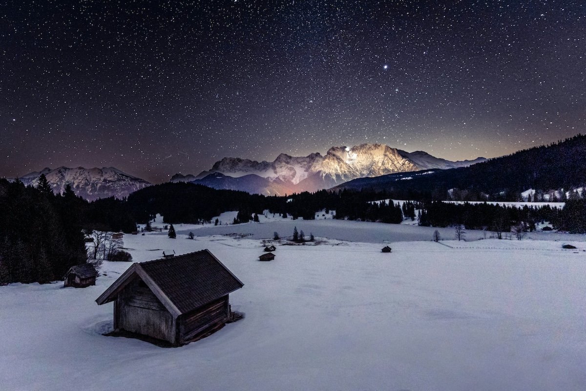 Night sky time-lapse photo with a cabin by a lake an mountain as the sun sets
