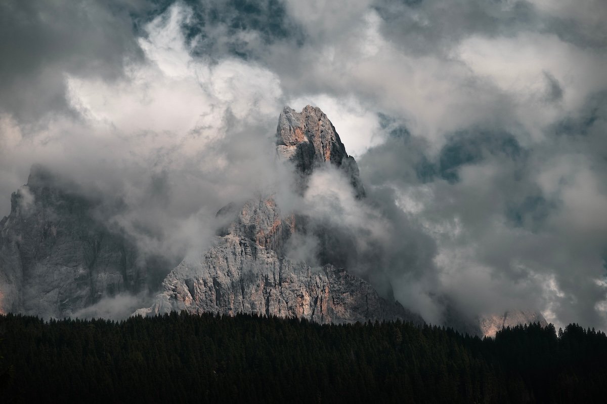 A sharp landscape photo of a mountain shrouded by clouds over trees