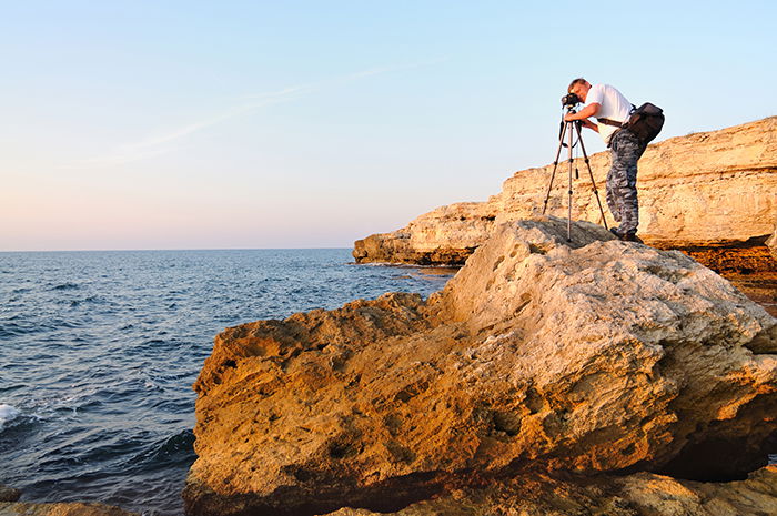 Man standing on rock and taking photo with tripod in blue hour.