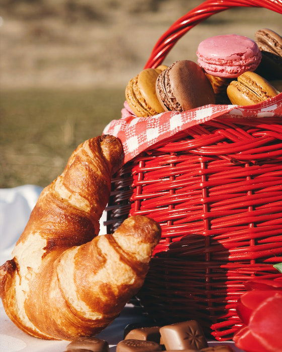 A close up of a red picnic basket full of macaroons and surrounded by croissants and chocolates