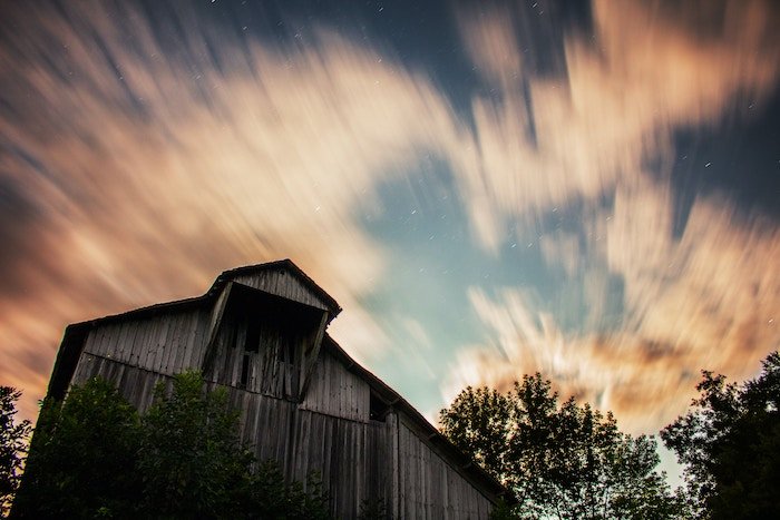 Long-exposure of the top of a barn, trees, and clouds in a twilight sky