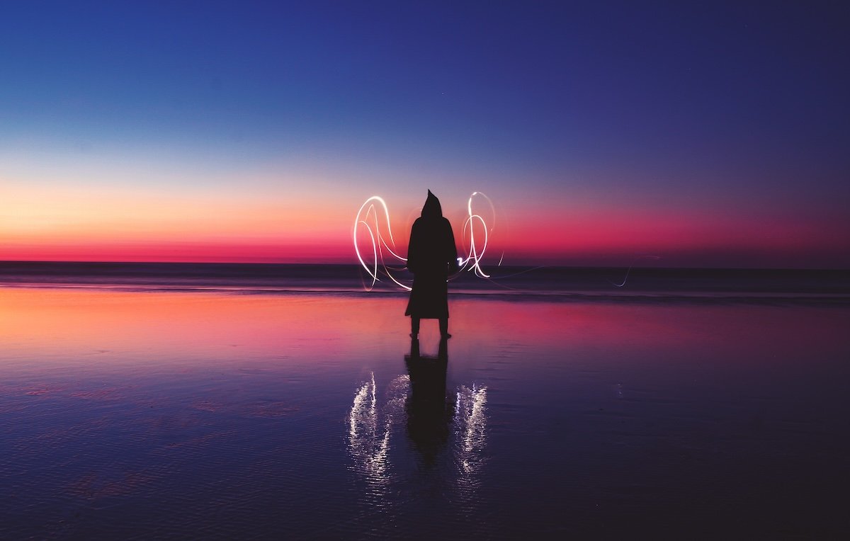 A person silhouetted on a beach at dusk with colorful sky and reflected ground with light graffiti painted in the air
