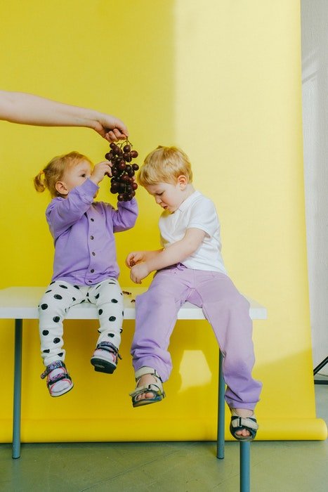Two kids sitting on a kitchen table eating grapes