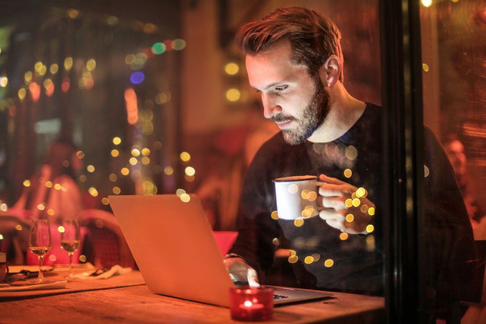 Uma imagem de um homem sentado em seu computador segurando uma xícara de café disparada por uma janela à noite