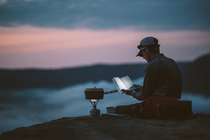 A hiker reading a book outside at dusk on a mountain ridge with a camp stove beside him