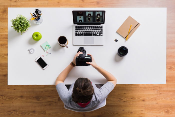 A woman from above, with a camera in her hands, and with a laptop, a mobile, an apple and coffee on the office table