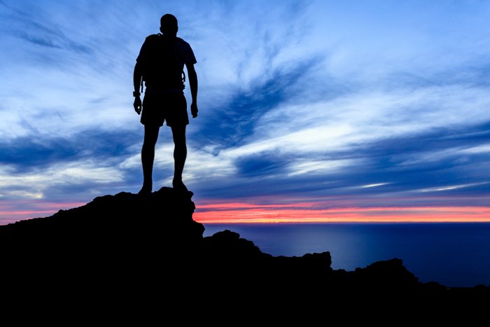 Man hiking silhouette in mountains, sunset and ocean over beautiful blue sky. Male hiker walking on top of mountain looking at beauty night landscape.