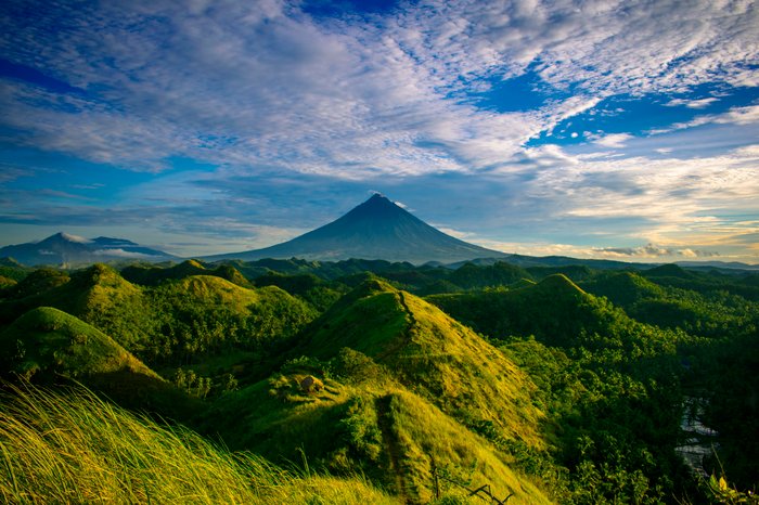 an image of a mountain in the background with green hills in the foreground and blue sky and clouds above