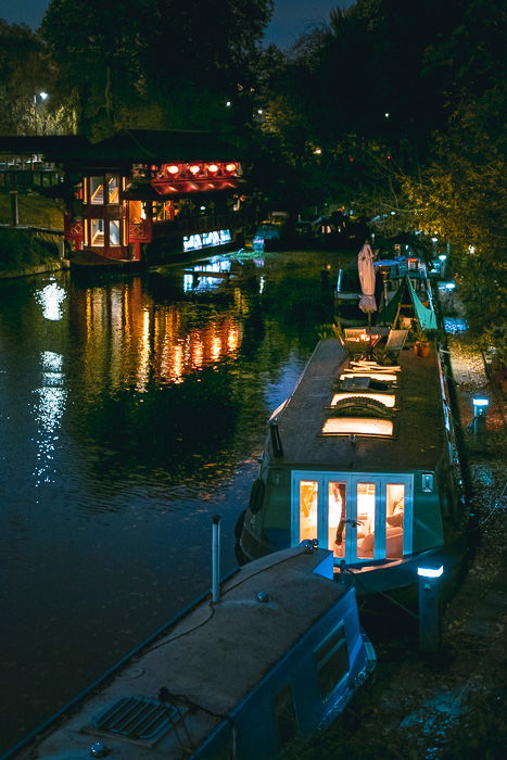 A nighttime photograph of an urban waterway with multiple boats, docked next to a riverbank. 