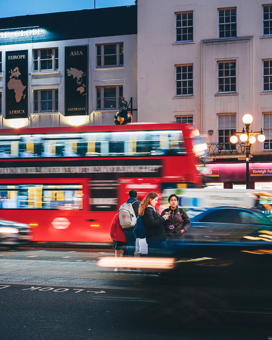 A night scene with a double-decker bus driving on the street, and people walking on the sidewalk. 