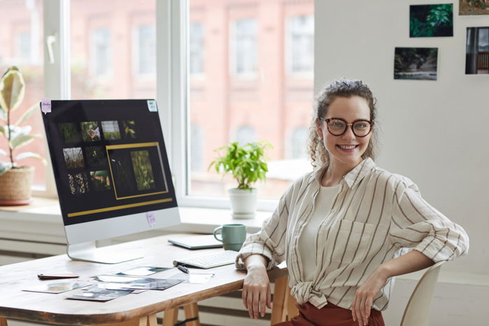 Portrait of young female photographer smiling at camera while posing at desk with photo editing software on computer screen