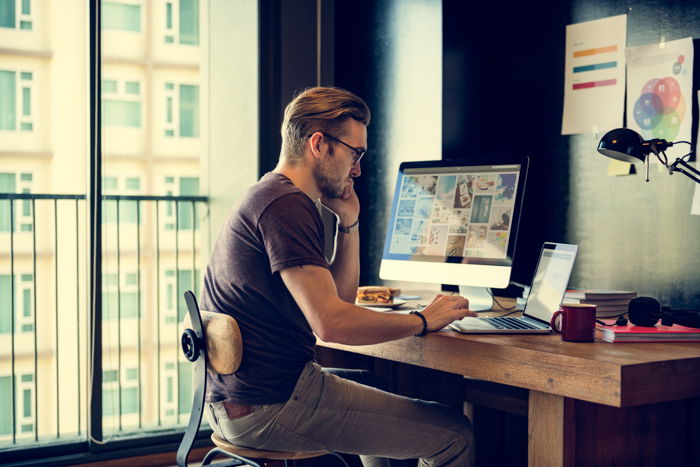 an image of a man in glasses working at a desk with desktop and laptop