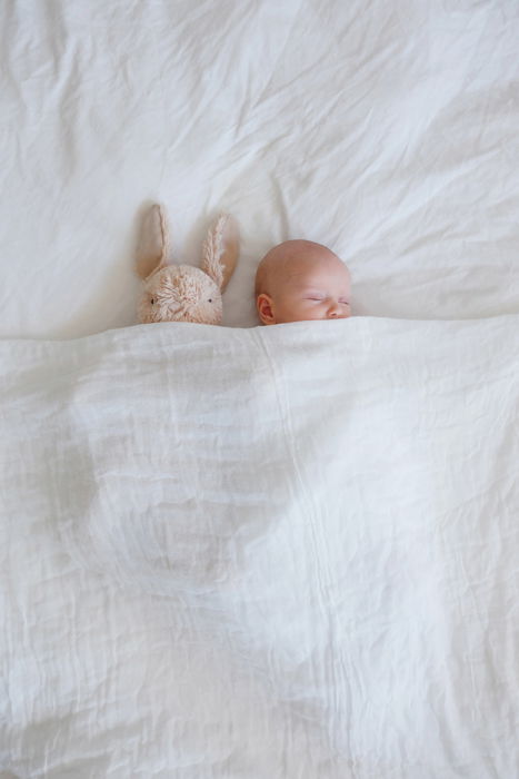 A baby in a crib with white sheets, surrounded by blankets, with a stuffed bunny on top. 