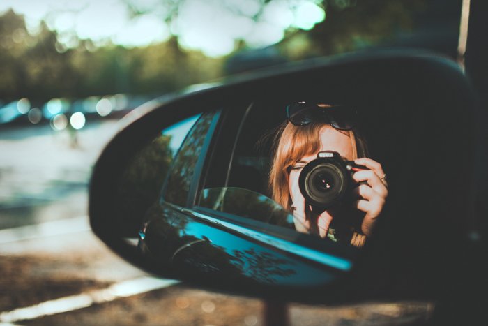 a side mirror reflection of a woman using a DSLR in the backseat of a car