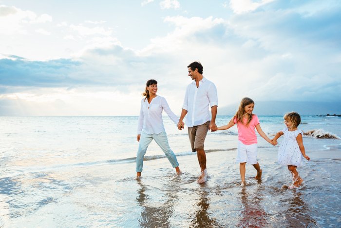 a family of four walks across the beach with their feet in the shallow water