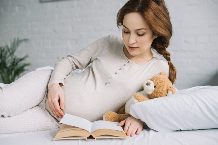 a pregnant woman poses on her side while reading a book and holding a teddy bear