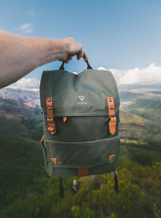 Product photography image of a backpack held up against a blurred natural landscape of sky, hills, and forest