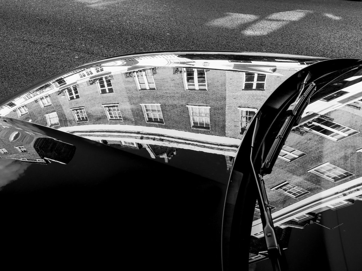 Black-and-white reflection of buildings in the hood and front mirror of a car parked in a street