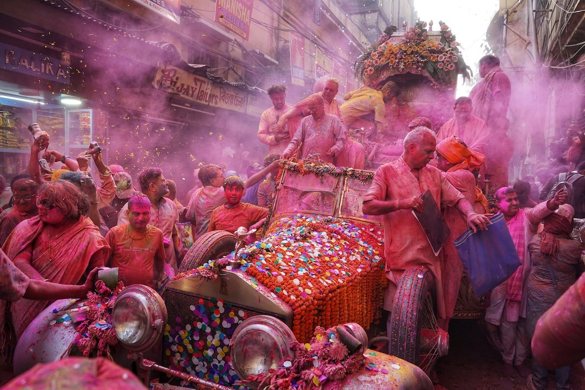 Street photo of a vintage car, people, and colorful confetti and powder during Holi celebration in India