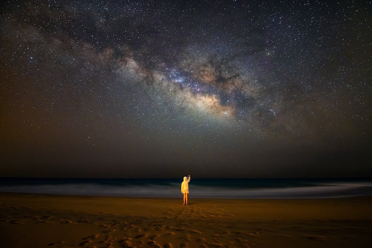 A person in a yellow coat standing on a beach under a starry night sky