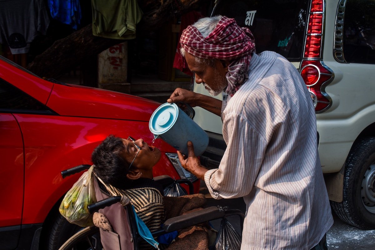 A man pouring water with a pitcher into the mouth of a boy in a wheelchair by parked cars