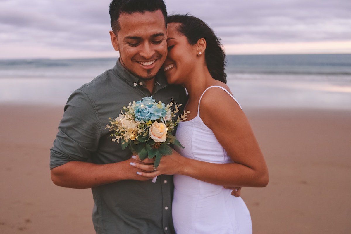 A wedding couple embracing and holding flowers on a beach