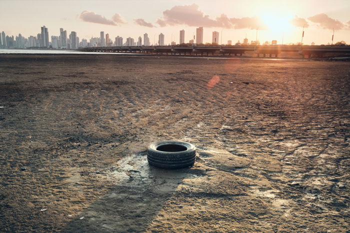 A documentary image of a rubber tyre on parched soil against an urban skyline