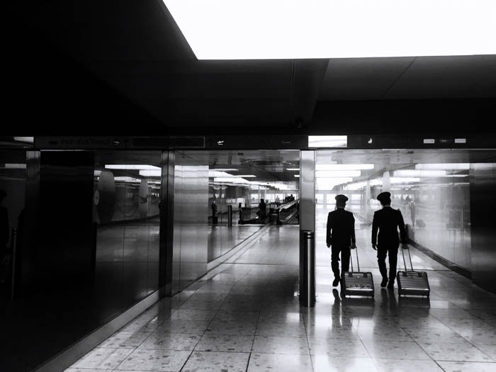 Candid shot of two pilots in an airport corridor. Black and white retro style image showing a back view of the pilots with their luggage, heading towards their flight.