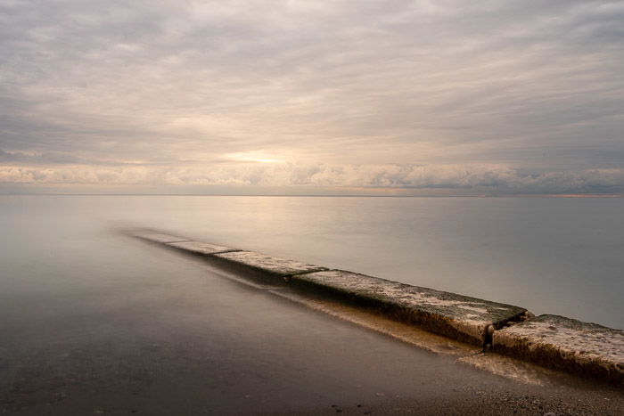 A landscape featuring a long pier jutting out from the shore into a large body of water under an overcast sky.