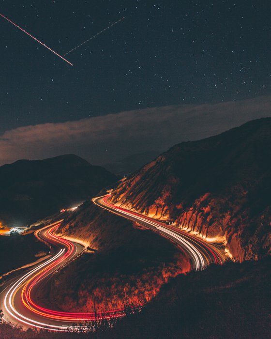 a time lapse photo of cars driving on a curved mountain road with shooting stars in the sky
