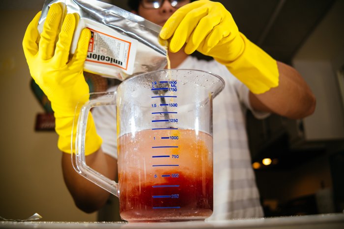 an image of a man in yellow gloves mixing film development chemicals in a glass measuring cup